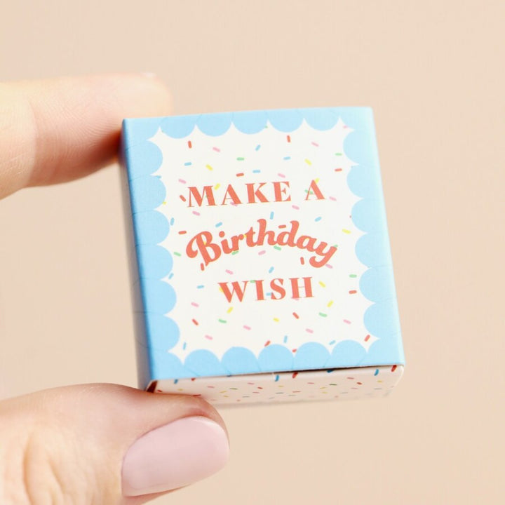 A small ceramic cupcake in a blue matchbox. The cupcake is frosted with white icing and has a red cherry on top. The matchbox has the text 