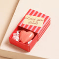 A small, pink heart-shaped object inside a red matchbox-sized gift box. The box has the text "Love You Loads" printed on it.