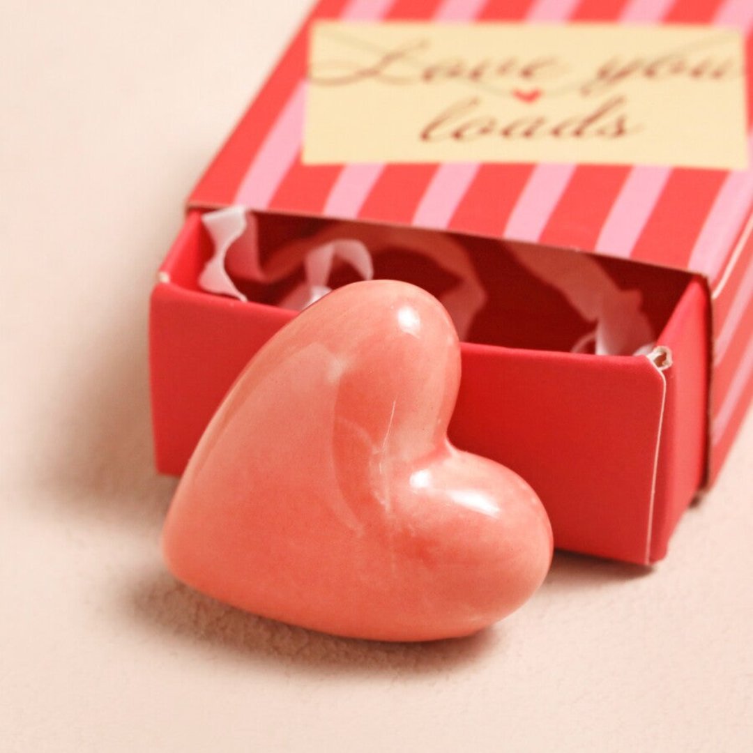 A small, pink heart-shaped object inside a red matchbox-sized gift box. The box has the text "Love You Loads" printed on it.