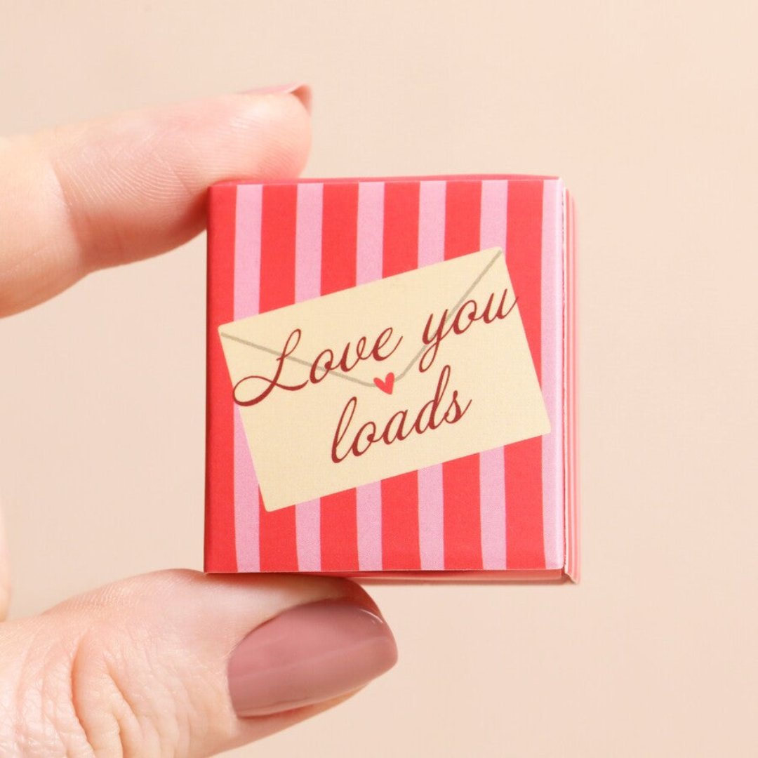 A small, pink heart-shaped object inside a red matchbox-sized gift box. The box has the text "Love You Loads" printed on it.