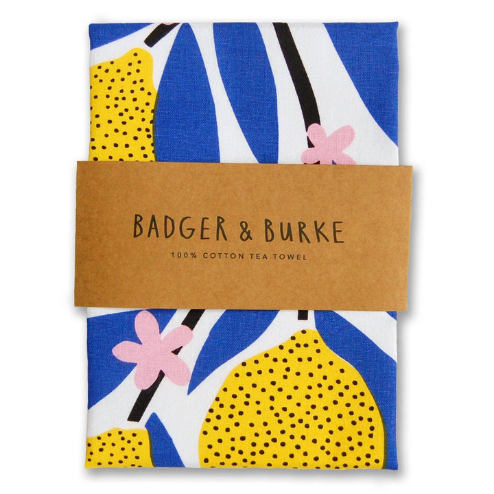 A white cotton tea towel with bright yellow lemons, blue leaves and pale pink flowers is folded in brown packaging that says Badger & Burke, 100% cotton tea towel.