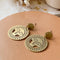 Brass Viviette Earrings. These elegant earrings feature circular brass discs with a sunburst cutout design and a small hammered brass circle at the top. The earrings are displayed on a soft, neutral-toned surface, adding a warm and sophisticated touch.