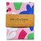 A pale pink cotton tea towel with bright blue, green, fuchsia, white and black geometric shapes is folded into a brown package. The packaging says Badger & Burke, 100% cotton tea towel.