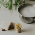 A bamboo bristle brush next to a cedar soap dish with soap on top and a pot filled with bubbles in front of a green plant.