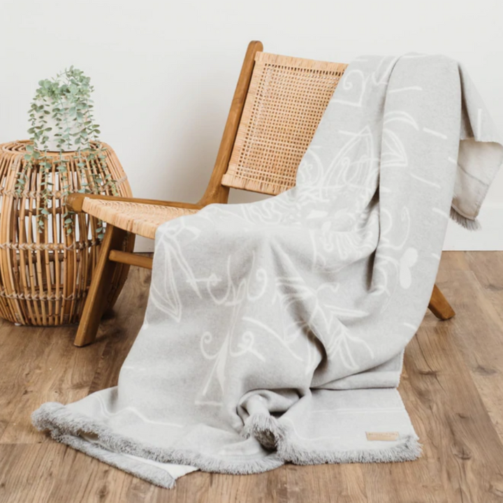 The Birch Bark Reversible Blanket by Mini Tipi, featuring a delicate white birch bark-inspired pattern on a grey background, draped over wooden chair. This reversible blanket is available in Ottawa, Ontario, Canada.