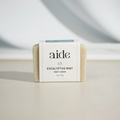 A bar of Aide Bodycare Eucalyptus Mint Soap, featuring a light beige color and a paper label with the 'aide' logo and 'Eucalyptus Mint' text, displayed on a light-colored surface with a shadow overlay. The soap is natural and handmade, available in Mississippi Mills, Ontario, Canada.