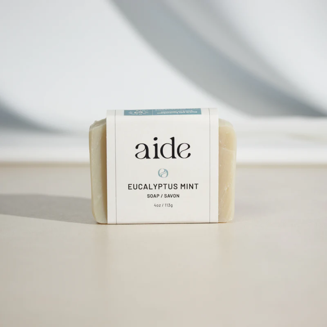 A bar of Aide Bodycare Eucalyptus Mint Soap, featuring a light beige color and a paper label with the 'aide' logo and 'Eucalyptus Mint' text, displayed on a light-colored surface with a shadow overlay. The soap is natural and handmade, available in Mississippi Mills, Ontario, Canada.