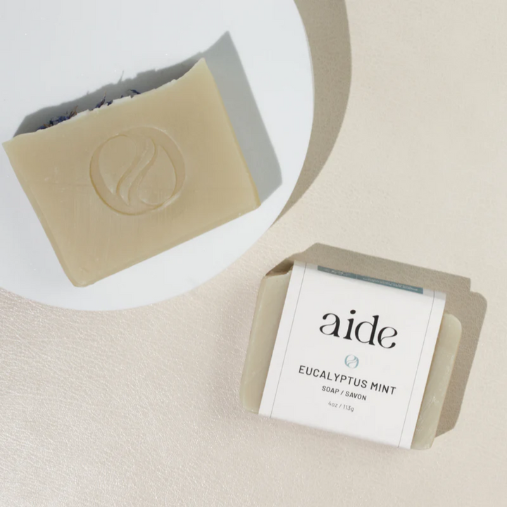 A bar of Aide Bodycare Eucalyptus Mint Soap, featuring a light beige color and a paper label with the 'aide' logo and 'Eucalyptus Mint' text, displayed on a light-colored surface with a shadow overlay. The soap is natural and handmade, available in Mississippi Mills, Ontario, Canada.