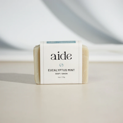 A bar of Aide Bodycare Eucalyptus Mint Soap, featuring a light beige color and a paper label with the 'aide' logo and 'Eucalyptus Mint' text, displayed on a light-colored surface with a shadow overlay. The soap is natural and handmade, available in Mississippi Mills, Ontario, Canada.