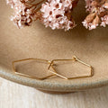 A pair of Agaveh Girl Gold Hexi Hoops, featuring a thin, gold wire hexagon design, displayed on a branded card with the text 'agaveh girl,' against a textured background with dried floral accents. The earrings are minimalist and geometric, available in Ottawa, Ontario, Canada.