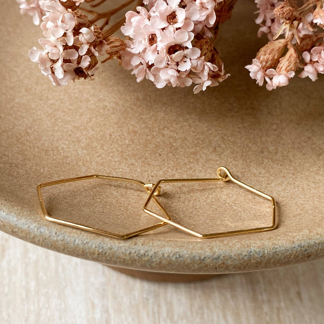 A pair of Agaveh Girl Gold Hexi Hoops, featuring a thin, gold wire hexagon design, displayed on a branded card with the text 'agaveh girl,' against a textured background with dried floral accents. The earrings are minimalist and geometric, available in Ottawa, Ontario, Canada.