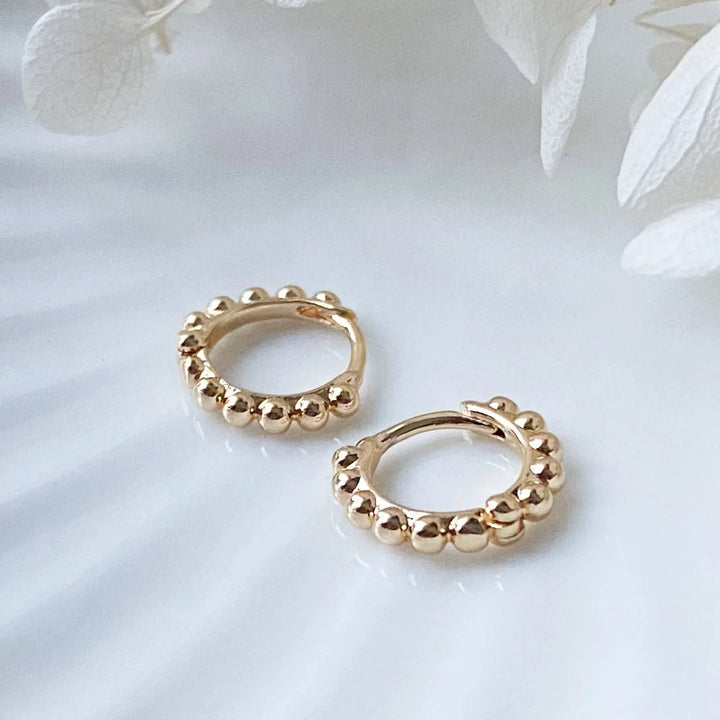 A pair of 14K Gold Ball Hoop Cuffs by agaveh girl, featuring a beaded design, displayed on a branded card with the text 'agaveh girl,' against a textured background. The earrings are dainty and high-quality, available in Ottawa, Ontario, Canada.