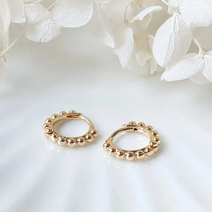A pair of 14K Gold Ball Hoop Cuffs by agaveh girl, featuring a beaded design, displayed on a branded card with the text 'agaveh girl,' against a textured background. The earrings are dainty and high-quality, available in Ottawa, Ontario, Canada.