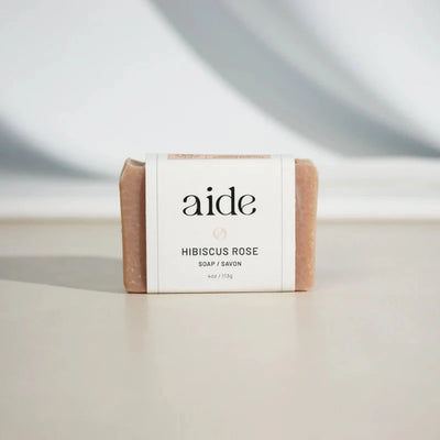 A bar of Aide Bodycare Hibiscus Rose Clay Soap, featuring a light pink color with a subtle texture and a paper label with the 'aide' logo and 'Hibiscus Rose' text, displayed on a light-colored surface with a shadow overlay. The soap is natural and handmade, available in Ottawa, Ontario, Canada.