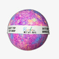 A multicolored bath bomb with purple, pink, and blue hues, packaged with a label that reads 'Rainbow NET WT. 150g'.