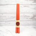 Two orange beeswax candles are standing upright on a white marble surface. They are tied together with a string and have a label that says "HONEY CANDLES 100% Pure Beeswax" and a honeycomb pattern.