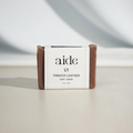 A bar of Aide Bodycare Tobacco Leather Soap, featuring a [describe color/pattern] and a paper label with the 'aide' logo and 'Tobacco Leather' text. The soap is natural and handmade, available in Mississippi Mills, Ontario, Canada.