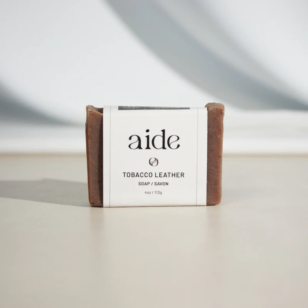 A bar of Aide Bodycare Tobacco Leather Soap, featuring a [describe color/pattern] and a paper label with the 'aide' logo and 'Tobacco Leather' text. The soap is natural and handmade, available in Mississippi Mills, Ontario, Canada.