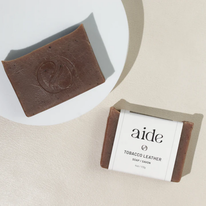 A bar of Aide Bodycare Tobacco Leather Soap, featuring a [describe color/pattern] and a paper label with the 'aide' logo and 'Tobacco Leather' text, . The soap is natural and handmade, available in Mississippi Mills, Ontario, Canada.