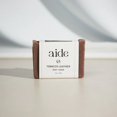 A bar of Aide Bodycare Tobacco Leather Soap, featuring a [describe color/pattern] and a paper label with the 'aide' logo and 'Tobacco Leather' text. The soap is natural and handmade, available in Mississippi Mills, Ontario, Canada.