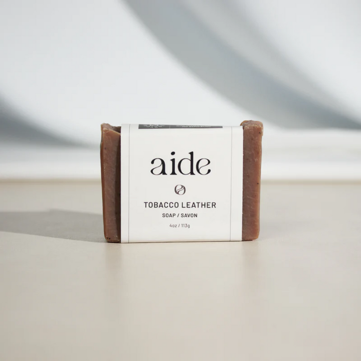 A bar of Aide Bodycare Tobacco Leather Soap, featuring a [describe color/pattern] and a paper label with the 'aide' logo and 'Tobacco Leather' text. The soap is natural and handmade, available in Mississippi Mills, Ontario, Canada.