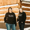 Two women in front of log house wearing black Be a nice Human crewneck sweatshirts