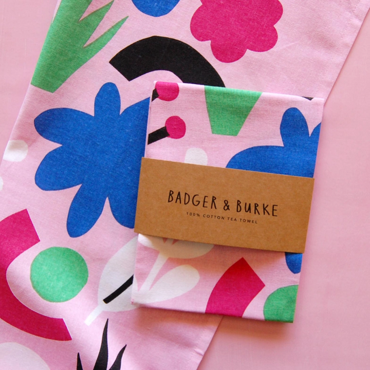 A pale pink cotton tea towel with blue, green, black and fuchsia geometric shapes lays open underneath a folded, packaged version of the same towel. The brown packaging says Badger & Burke, 100% cotton tea towel. 