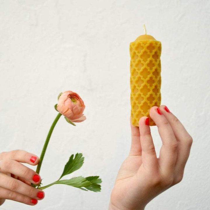 One hand holding a pink peony, and another hand holding up a yellow beeswax pillar candle with a design inspired by medieval windows