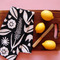 A black and white floral patterned, cotton tea towel lays folded on a wooden cutting board next to a knife and three lemons.